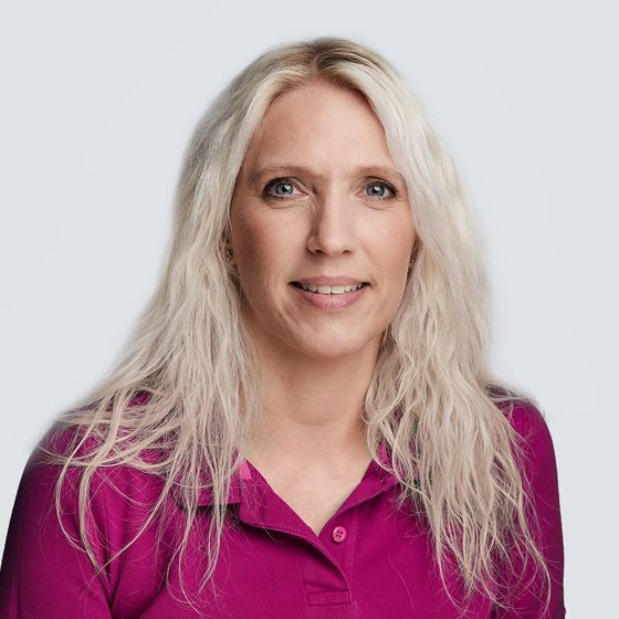 Frau mit langen blonden Haaren, die ein rosa Hemd mit Kragen trägt, lächelt vor einem weißen Hintergrund.