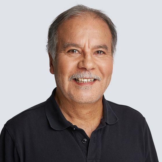 Ein Mann mit Schnurrbart lächelt in einem dunklen Hemd vor einem hellgrauen Hintergrund.