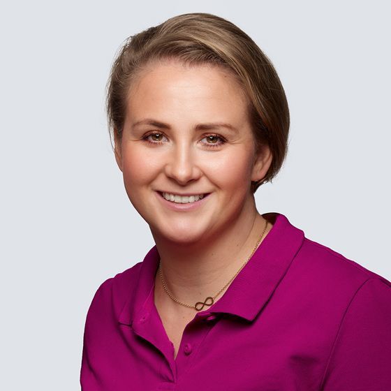 Frau mit kurzen blonden Haaren, die ein magentafarbenes Hemd trägt, lächelt und trägt eine goldene Halskette.