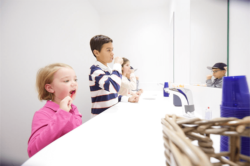 Kinder putzen sich an einem weißen Waschbecken die Zähne. Ein blondes Mädchen öffnet den Mund, während zwei andere sich vor einem Spiegel die Zähne putzen.
