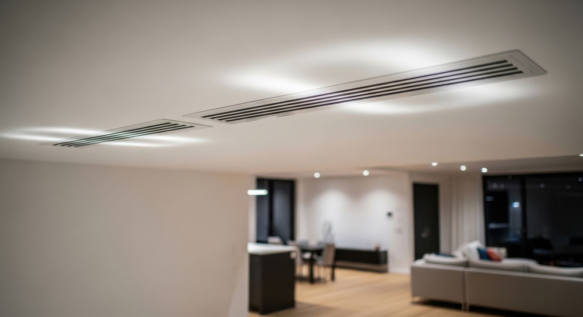 Salon minimaliste moderne avec des spots encastrés au plafond, des murs blancs et canapé en arrière-plan et au premier plan une VMC.