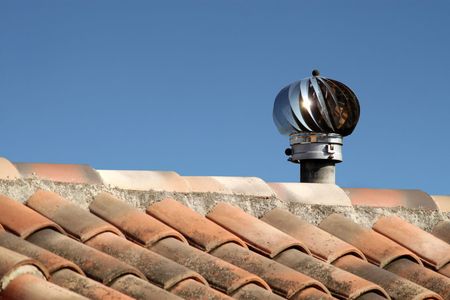 Des tuiles de toit avec une turbine de ventilation métallique se détachant sur un ciel bleu clair.