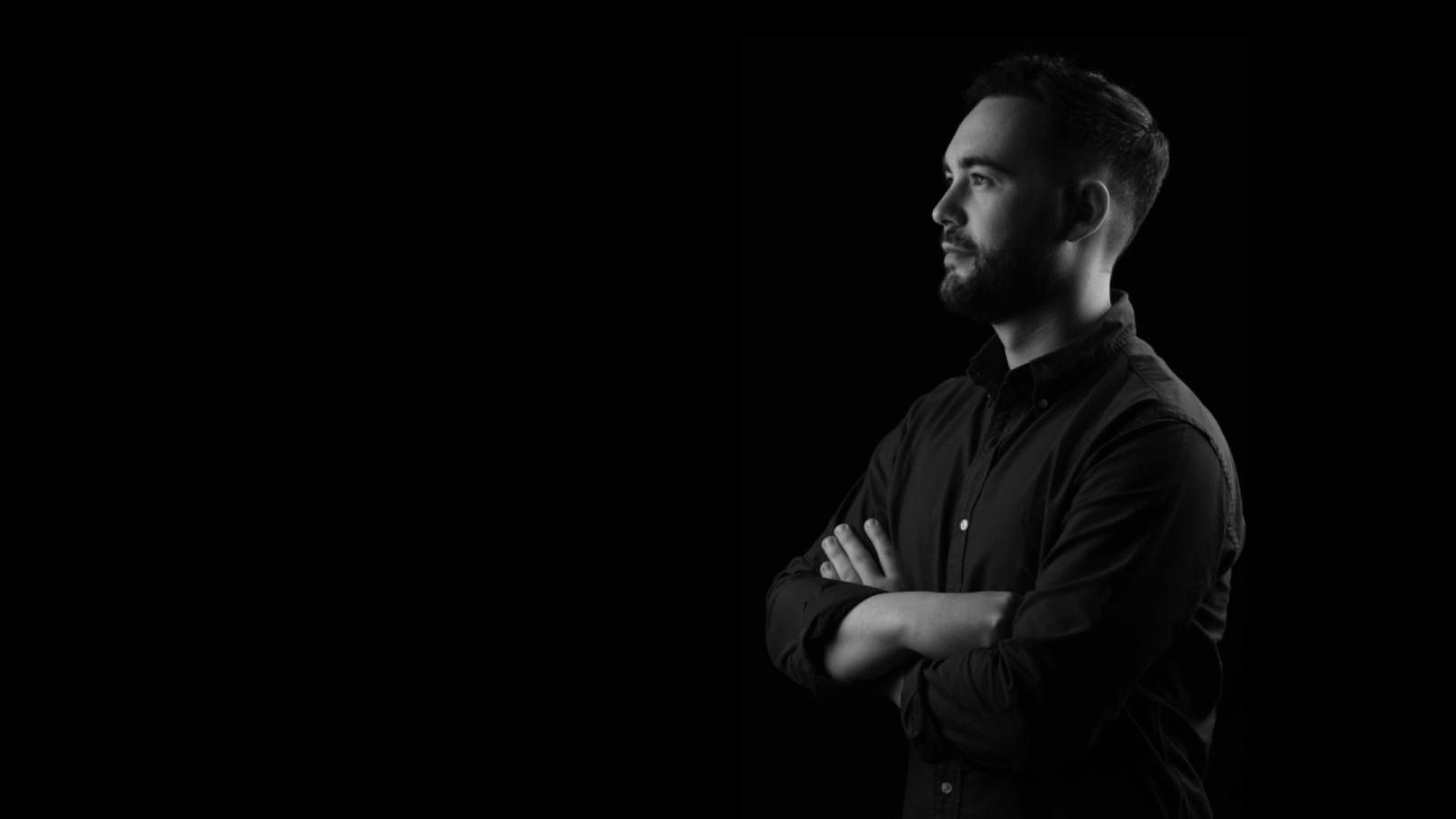 Un hombre con barba está de pie en la oscuridad con los brazos cruzados.