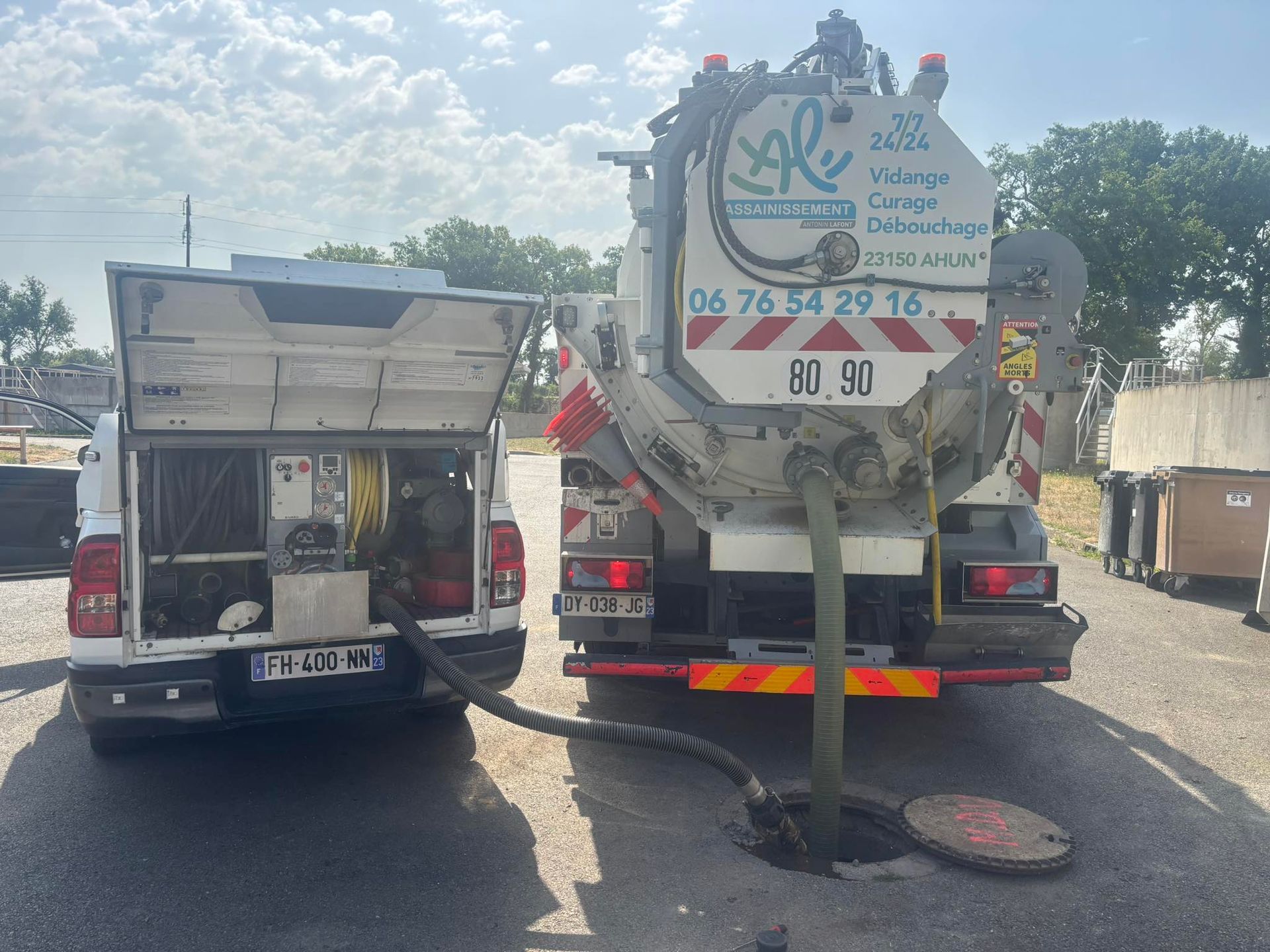 Camion en train d'aspirer un égout, relié par un tuyau. Camion blanc à gauche et camion aspirateur spécialisé à droite.