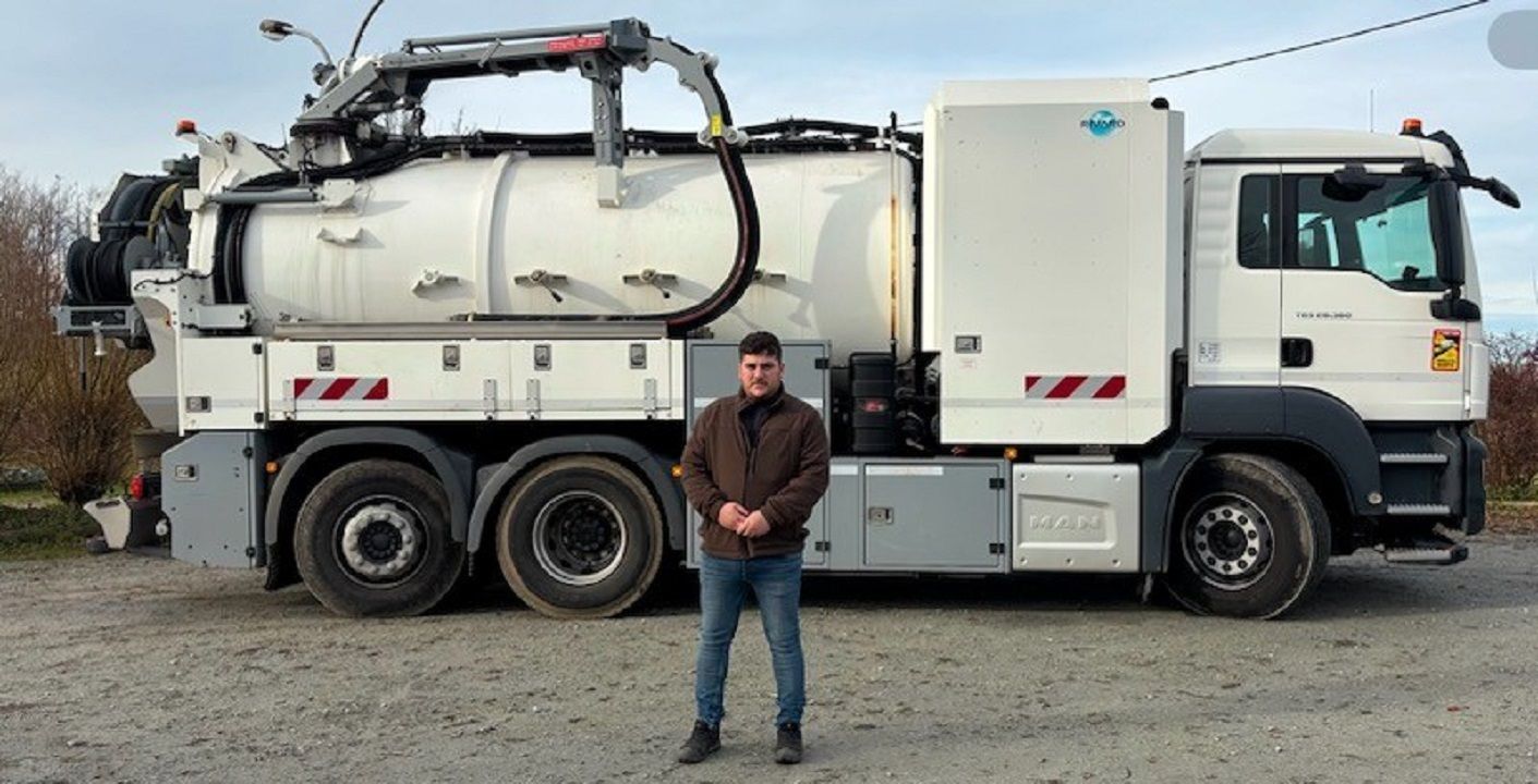 Un homme se tient devant un camion-citerne blanc auquel sont fixés un tuyau et un bras noirs.