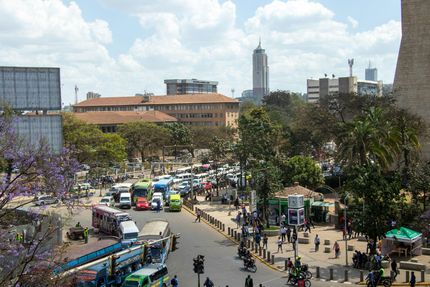 Busy city square with crowds, buses, and trees under a partly cloudy sky