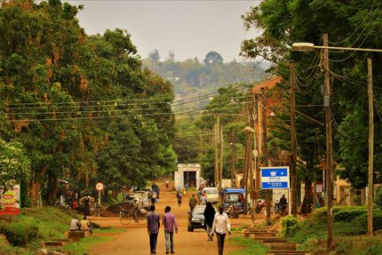 People walking on a dirt road in a green village with trees, power poles, and distant hills