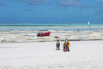 People walking on a bright white beach near a red boat, with blue water and sky in the background.