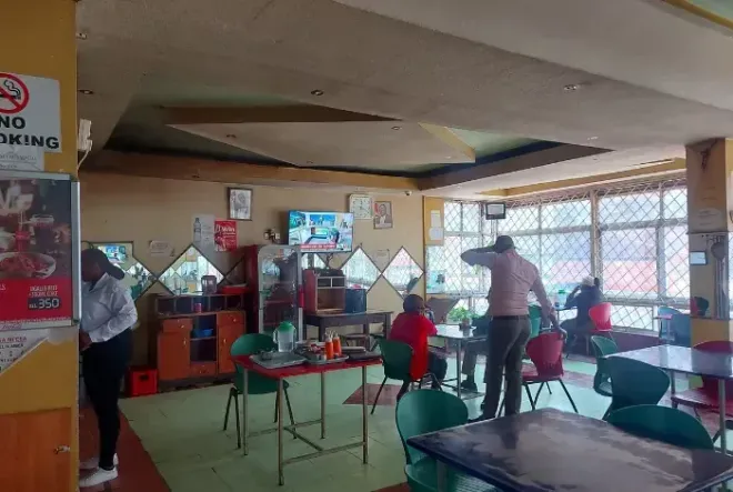 Small restaurant interior with red chairs, green tables, and people standing near the counter.