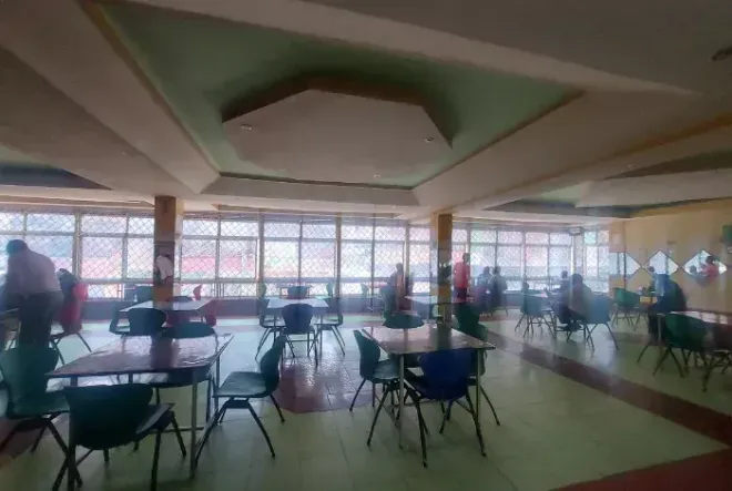 Spacious cafeteria with round tables and chairs by large windows, a few people seated and standing.
