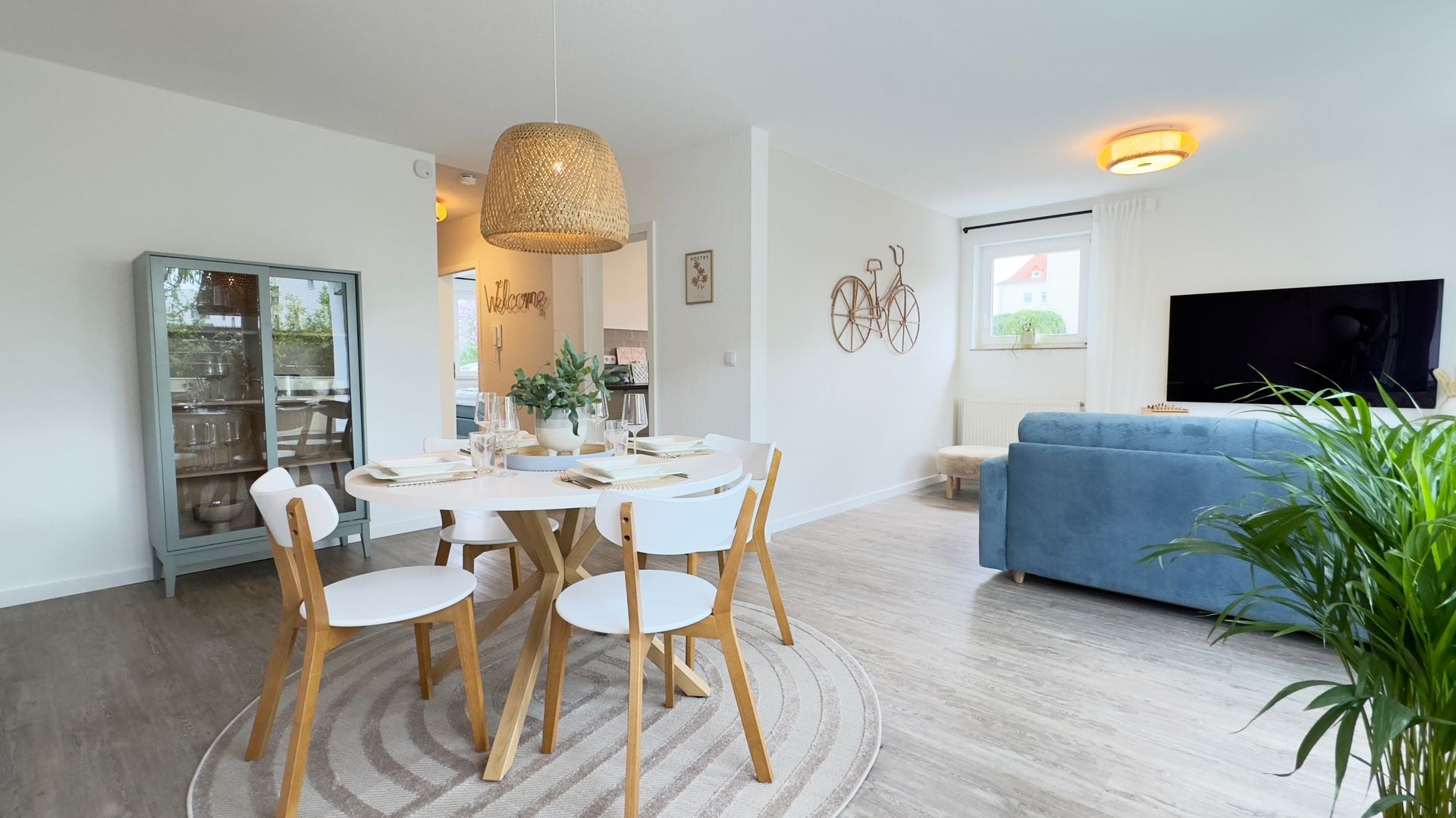 Wohnzimmer der Ferienwohnung Am Park mit rundem Esstisch, Stühlen und grüner Pflanze auf dem Tisch. Im Hintergrund ein blaues Sofa.