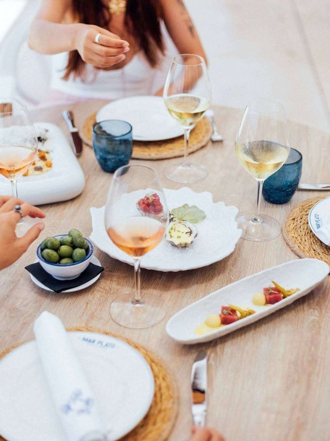 Gente cenando en una mesa soleada con copas de vino, ensaladas y aperitivos.