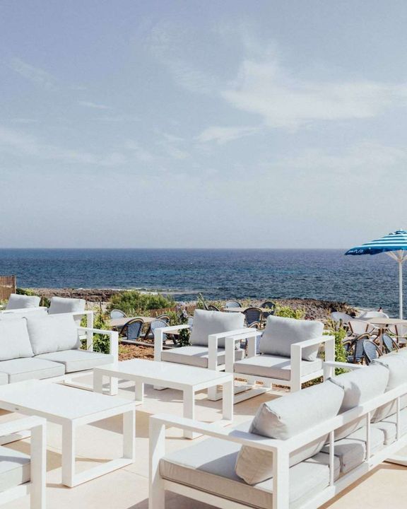 Tumbonas y mesas blancas en un patio junto al mar con vistas al océano bajo un cielo despejado.