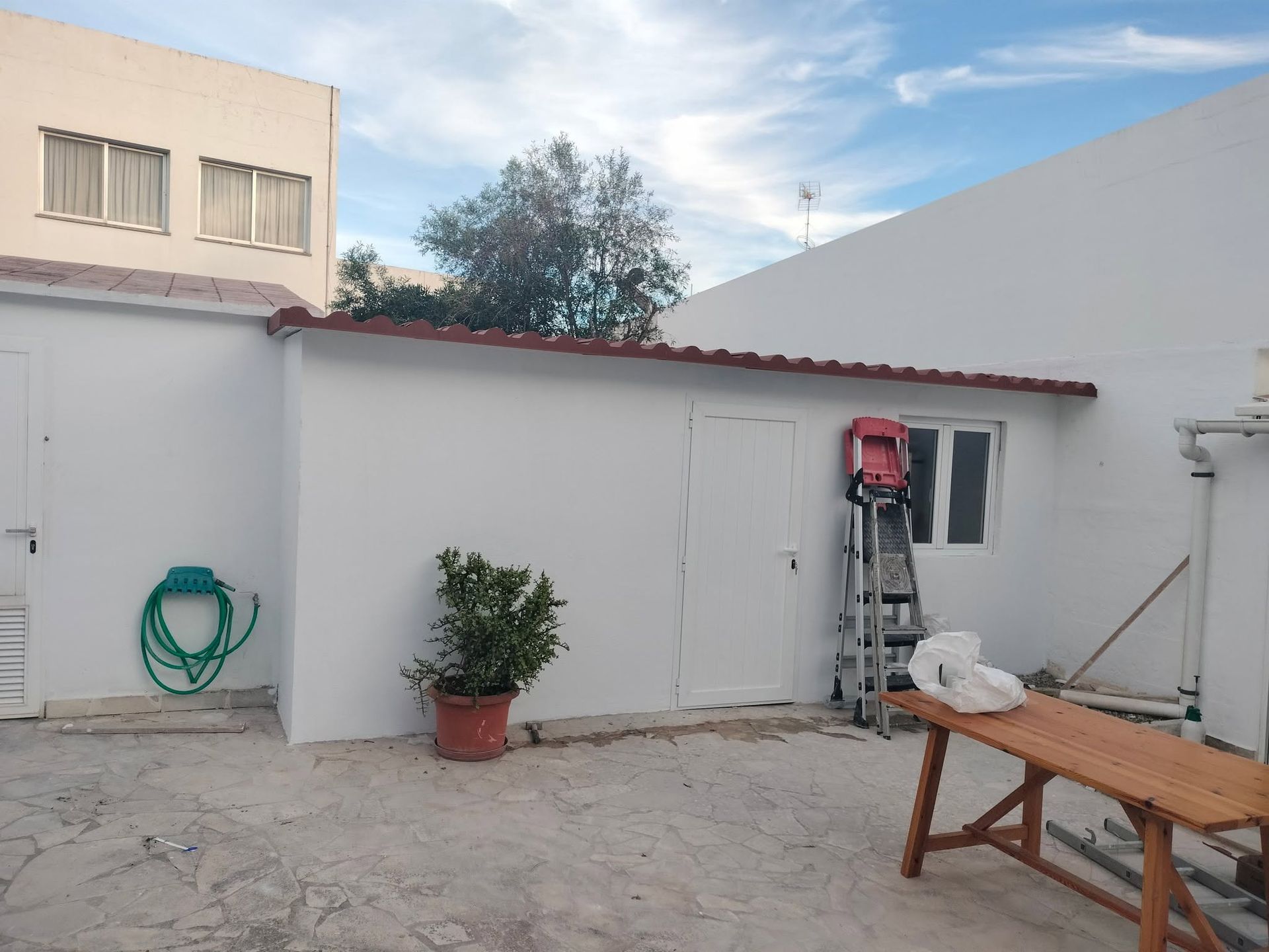 Pequeño patio con paredes blancas, una escalera, una planta en maceta, una manguera y una mesa de madera bajo un cielo azul.