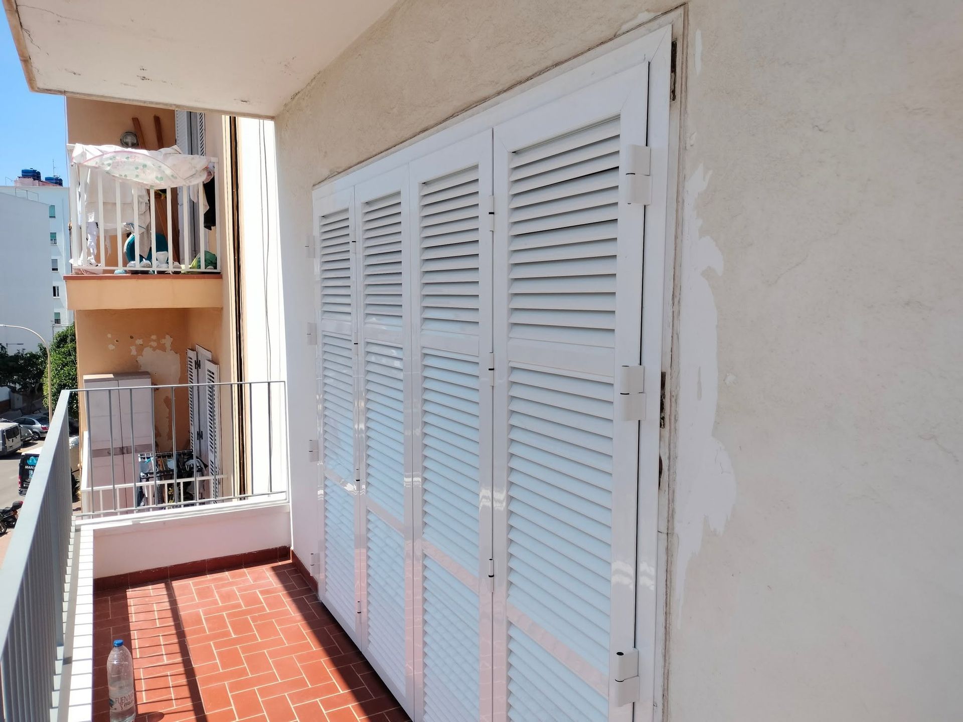 Balcón estrecho con puertas blancas contraventanas, suelo de baldosas rojas y edificios de apartamentos colindantes.