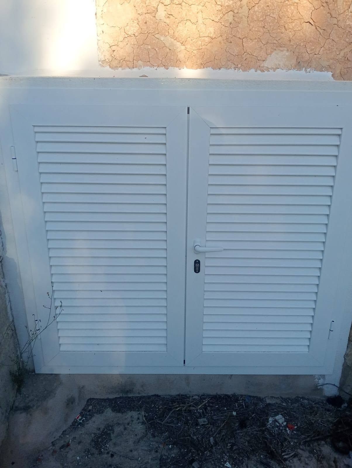 Puertas de servicio blancas con celosías, instaladas en una pared de estuco, con luz solar en la parte superior 