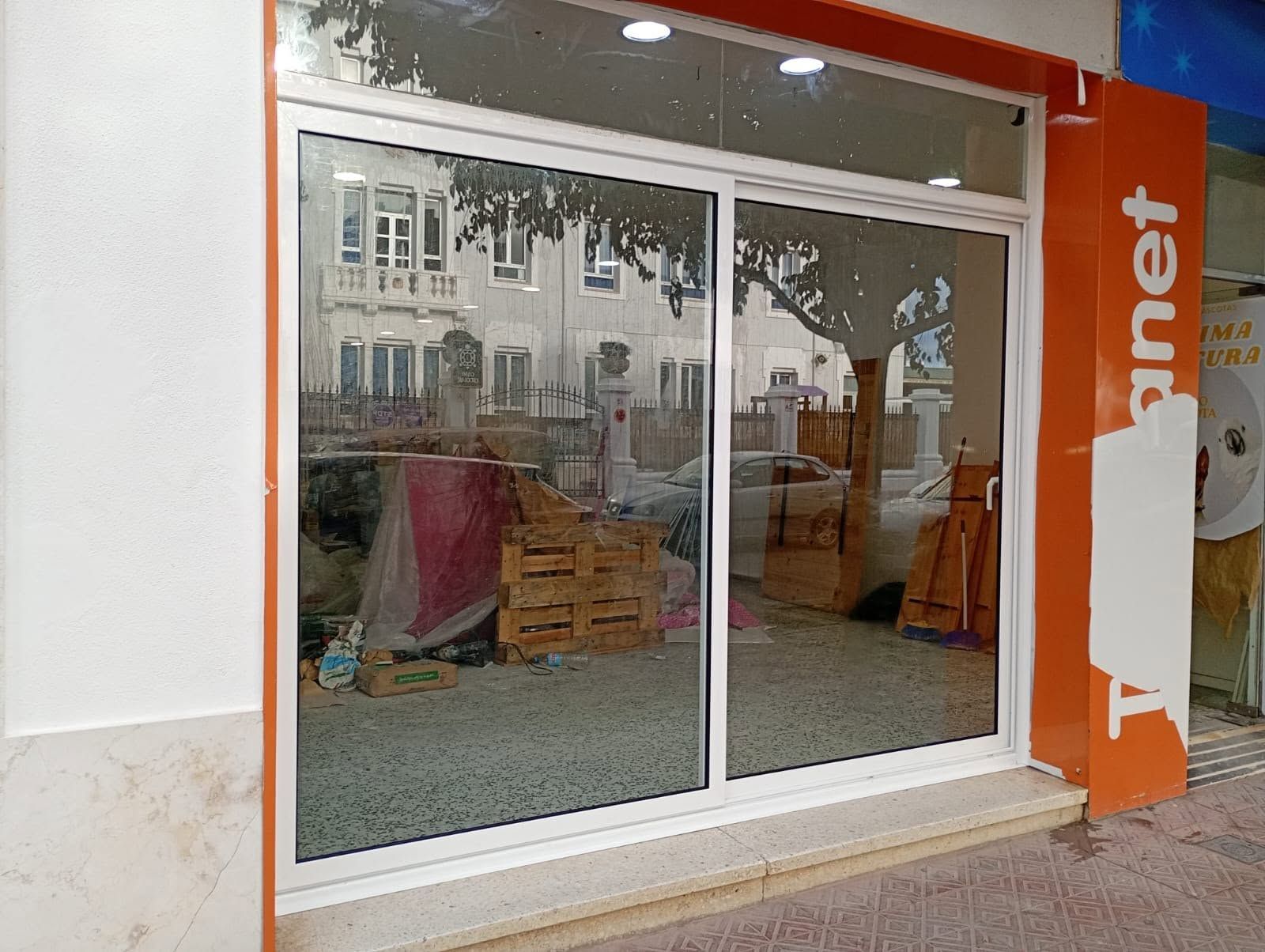 Escaparate cerrado con puerta corredera de cristal, marco naranja y desorden interior visible tras el reflejo.