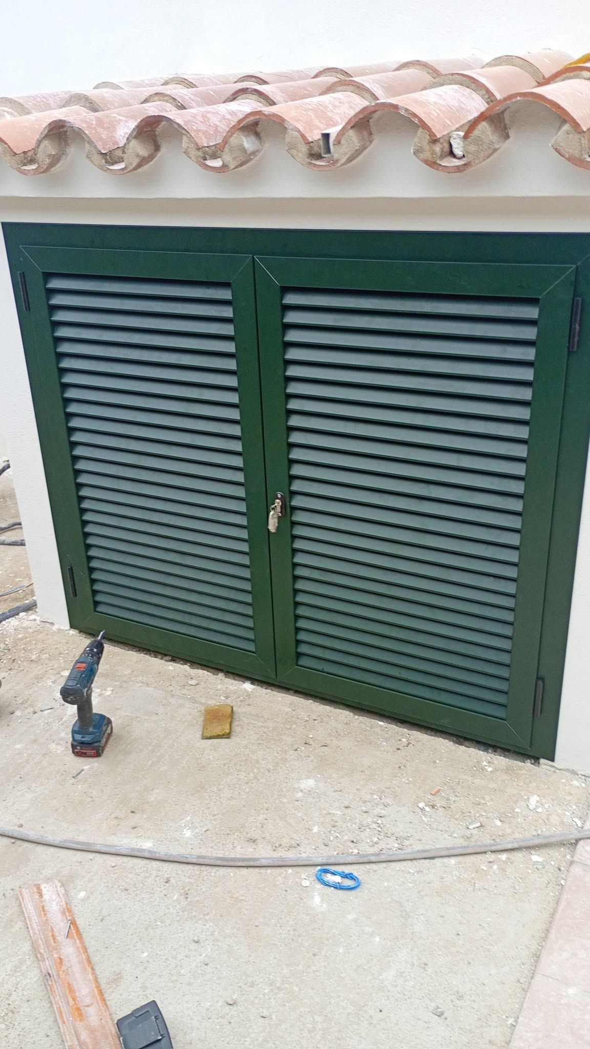 Puerta de armario verde con persianas bajo un techo de tejas en un patio de hormigón.