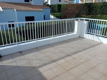 Terraza soleada con baldosas y barandilla blanca con vistas a las casas y la vegetación.