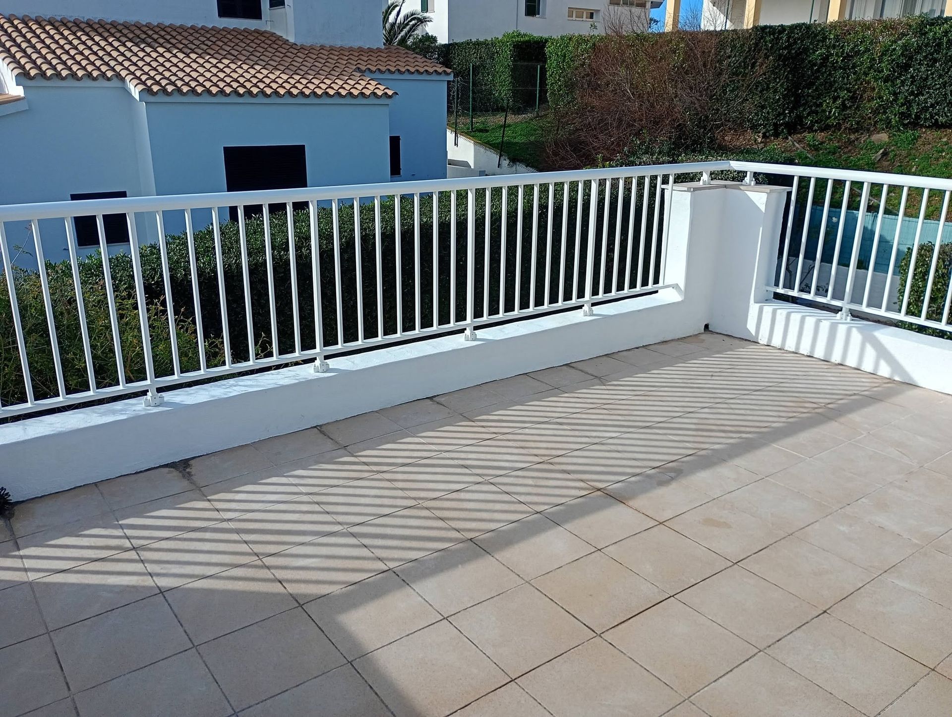 Terraza soleada con baldosas, barandilla blanca y casa azul al fondo.