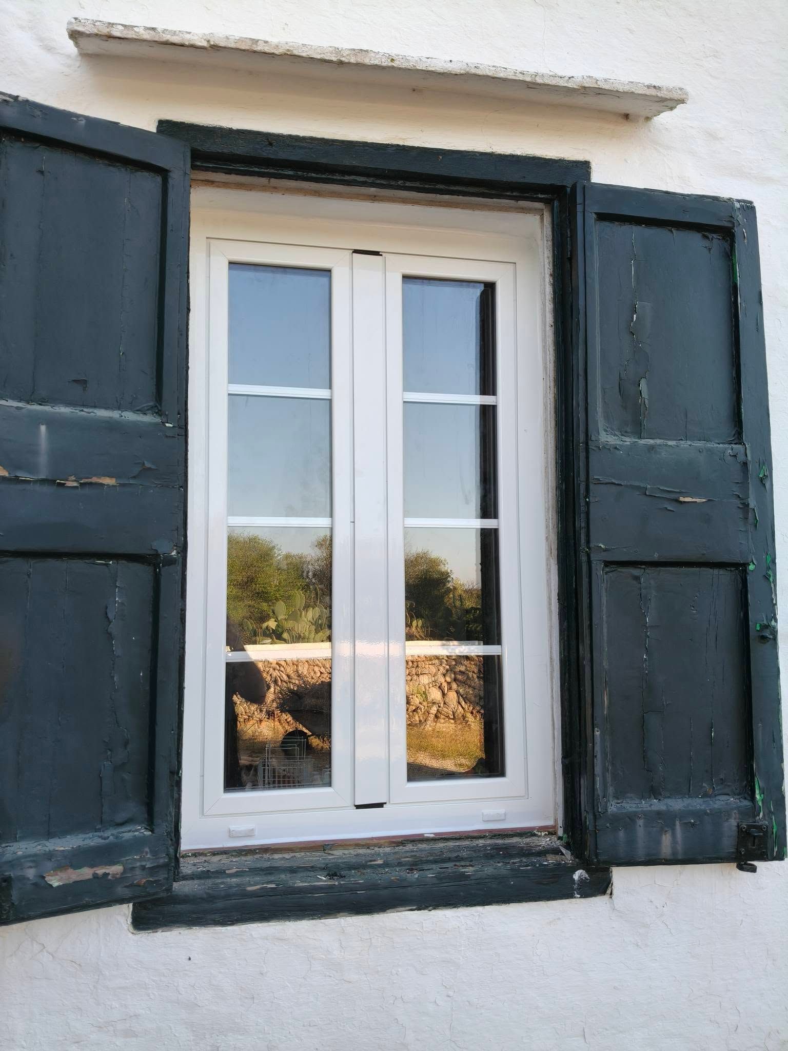 Ventana blanca con contraventanas azul oscuro sobre una pared blanca, que refleja los árboles y el paisaje exterior.