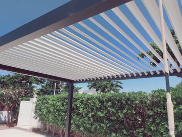 Patio trasero con pérgola a rayas, seto y armario blanco de almacenamiento bajo la luz del sol.