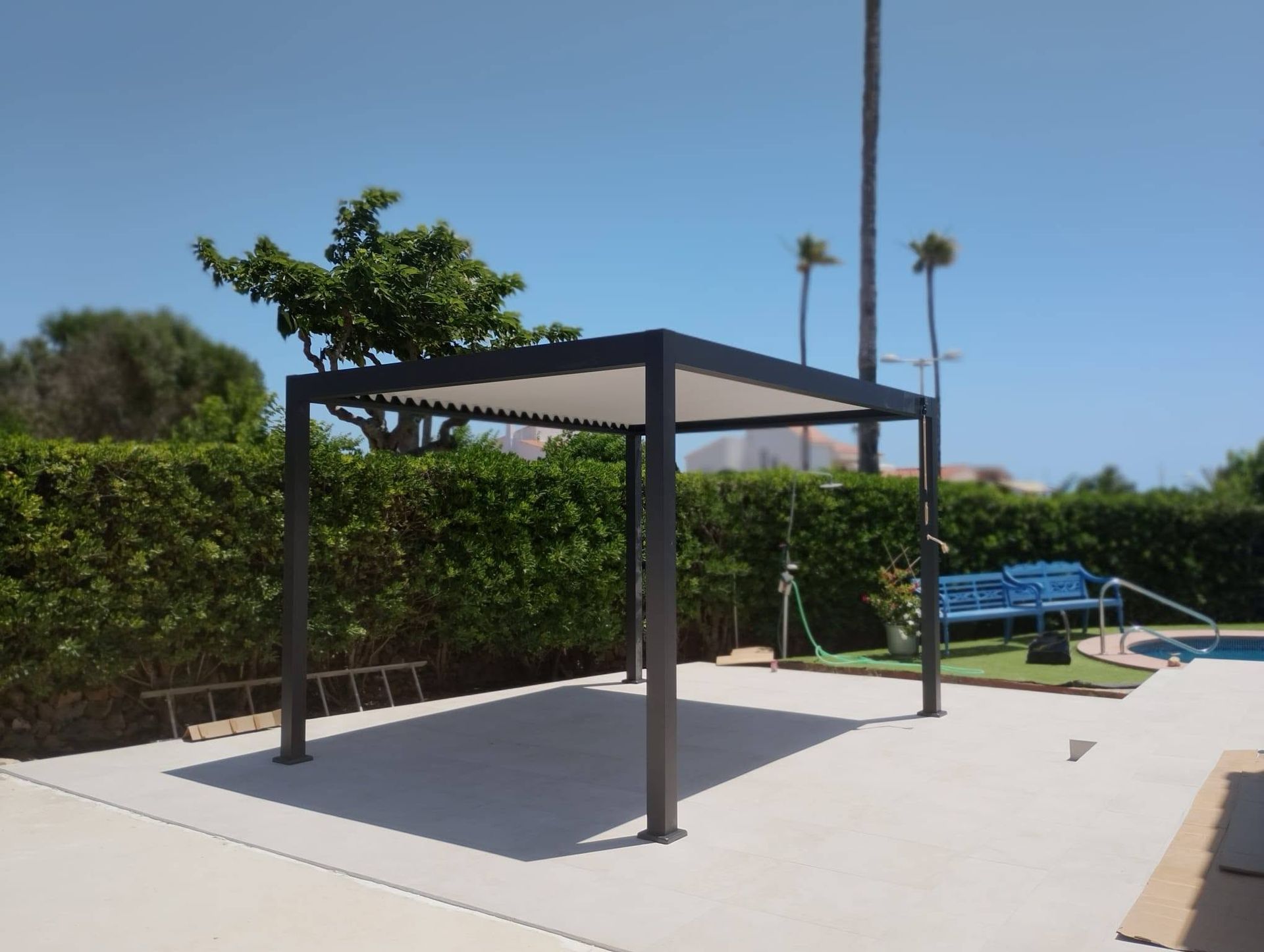 Patio exterior de hormigón con una pérgola de metal negro junto a un seto y un banco azul bajo un cielo despejado.
