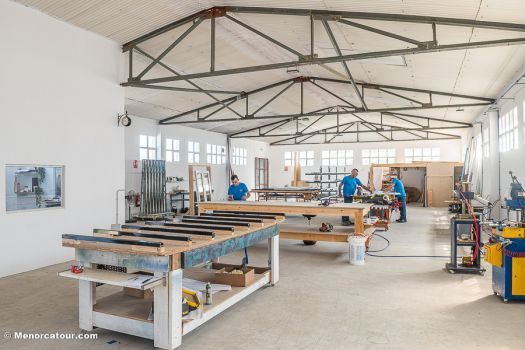 Taller luminoso con largas mesas de trabajo, herramientas y varias personas bajo un alto techo de cerchas metálicas.