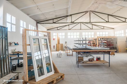 Espacio de trabajo tipo estudio luminoso con grandes ventanales, marcos de madera, mesas y herramientas bajo un techo alto.