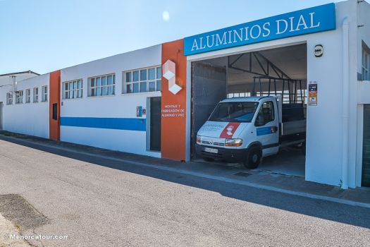 Edificio industrial blanco con detalles azules y un coche aparcado en la entrada bajo un letrero de 