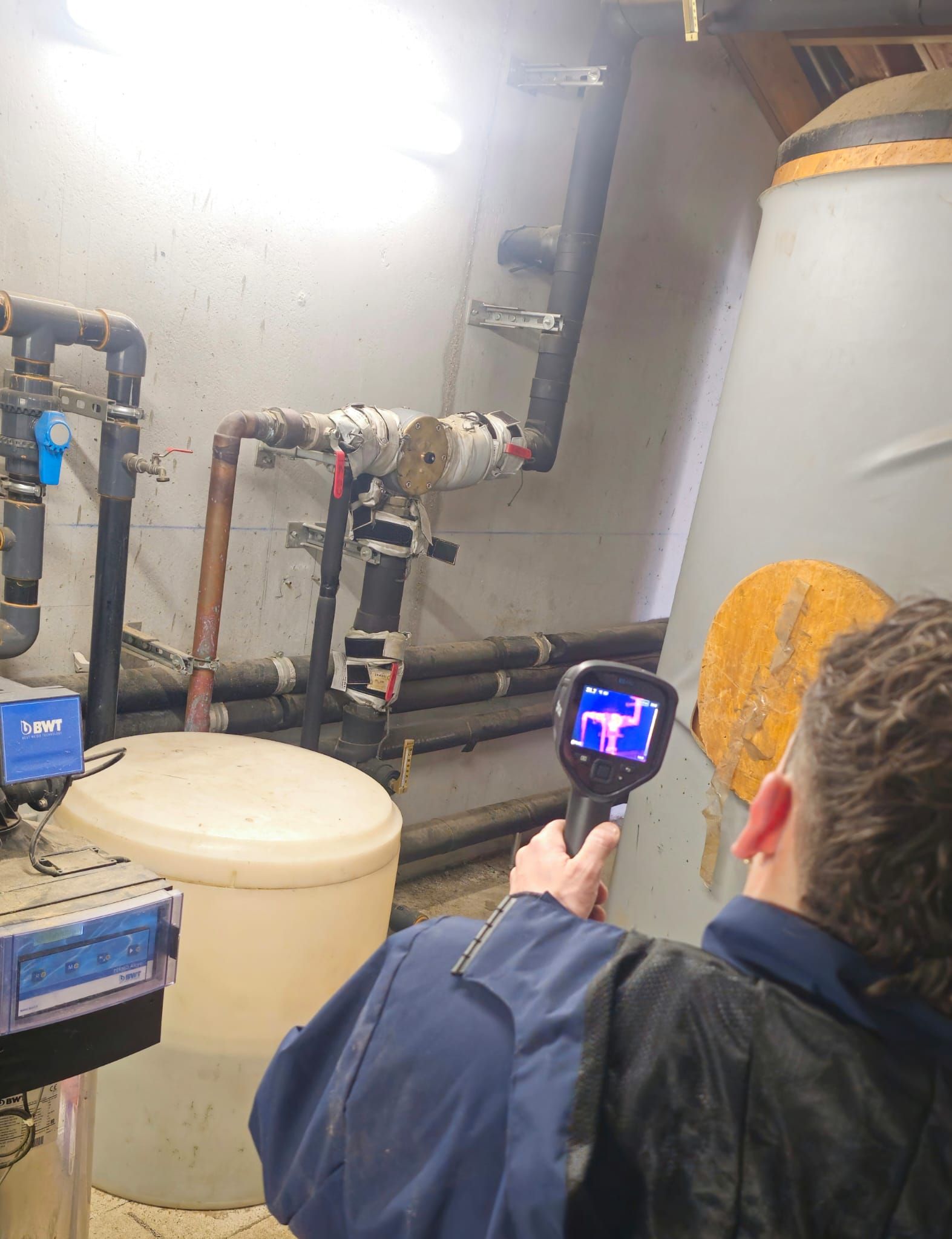 Une personne utilise une caméra thermique sur des tuyaux et des réservoirs dans un local technique ; la caméra révèle des signatures thermiques.