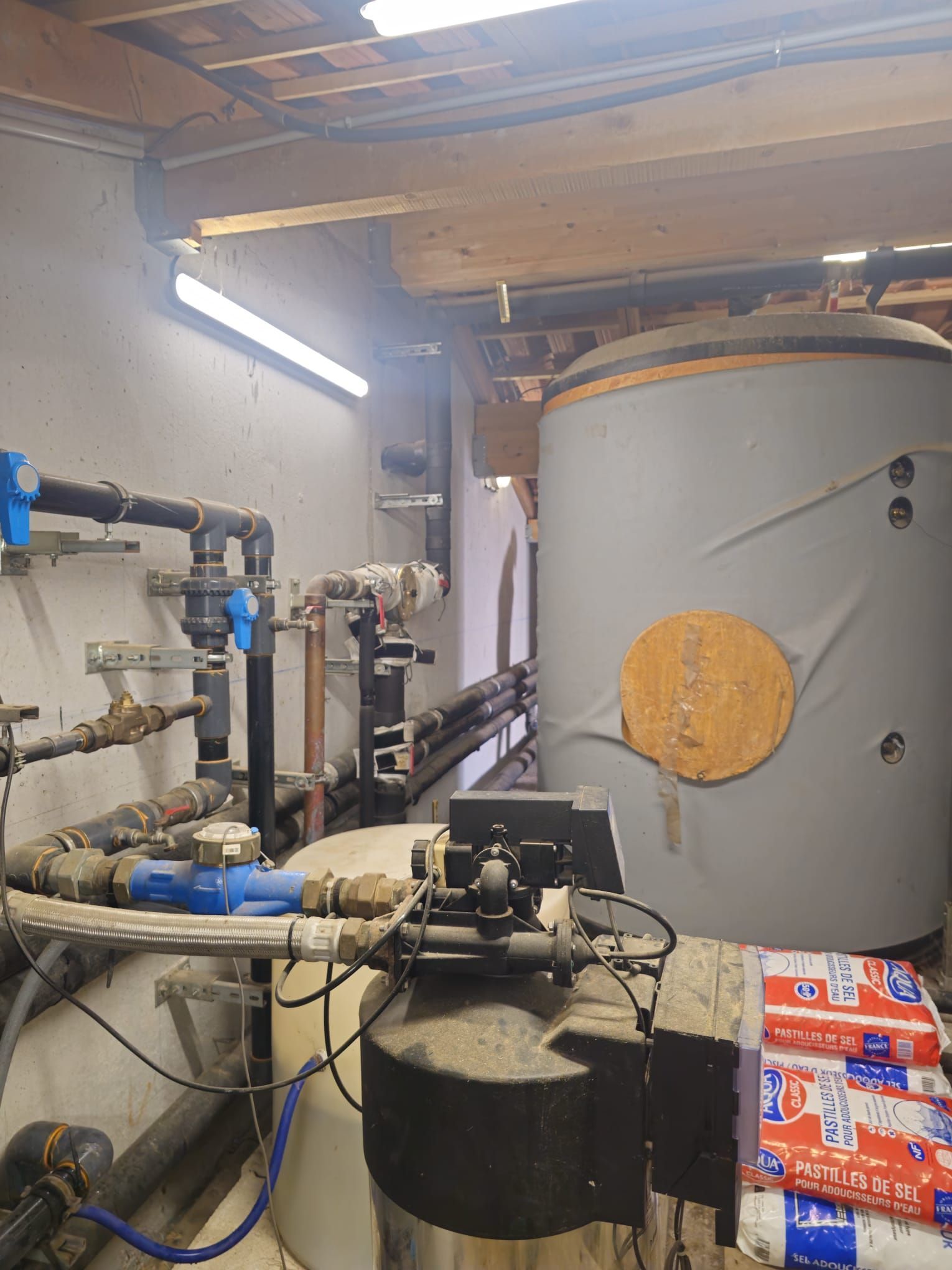 Local technique avec chauffe-eau et système de filtration ; tuyaux, vannes et grand réservoir visibles.