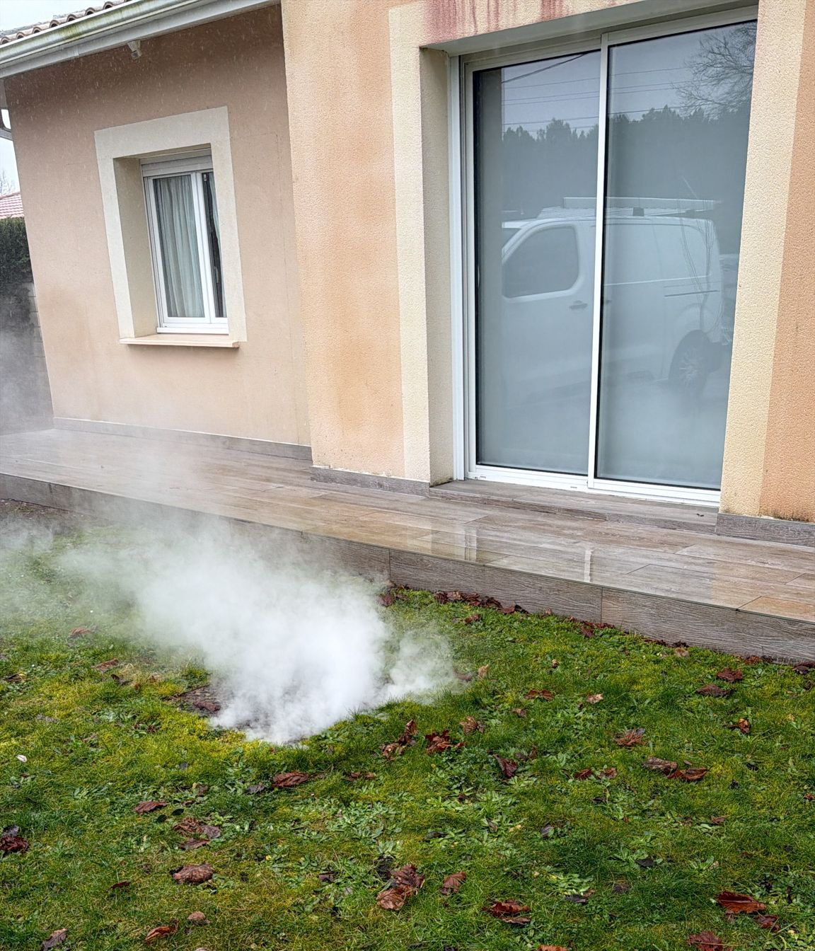 De la vapeur s'élève de l'herbe près d'une maison avec une porte coulissante en verre et un chemin de pierre.