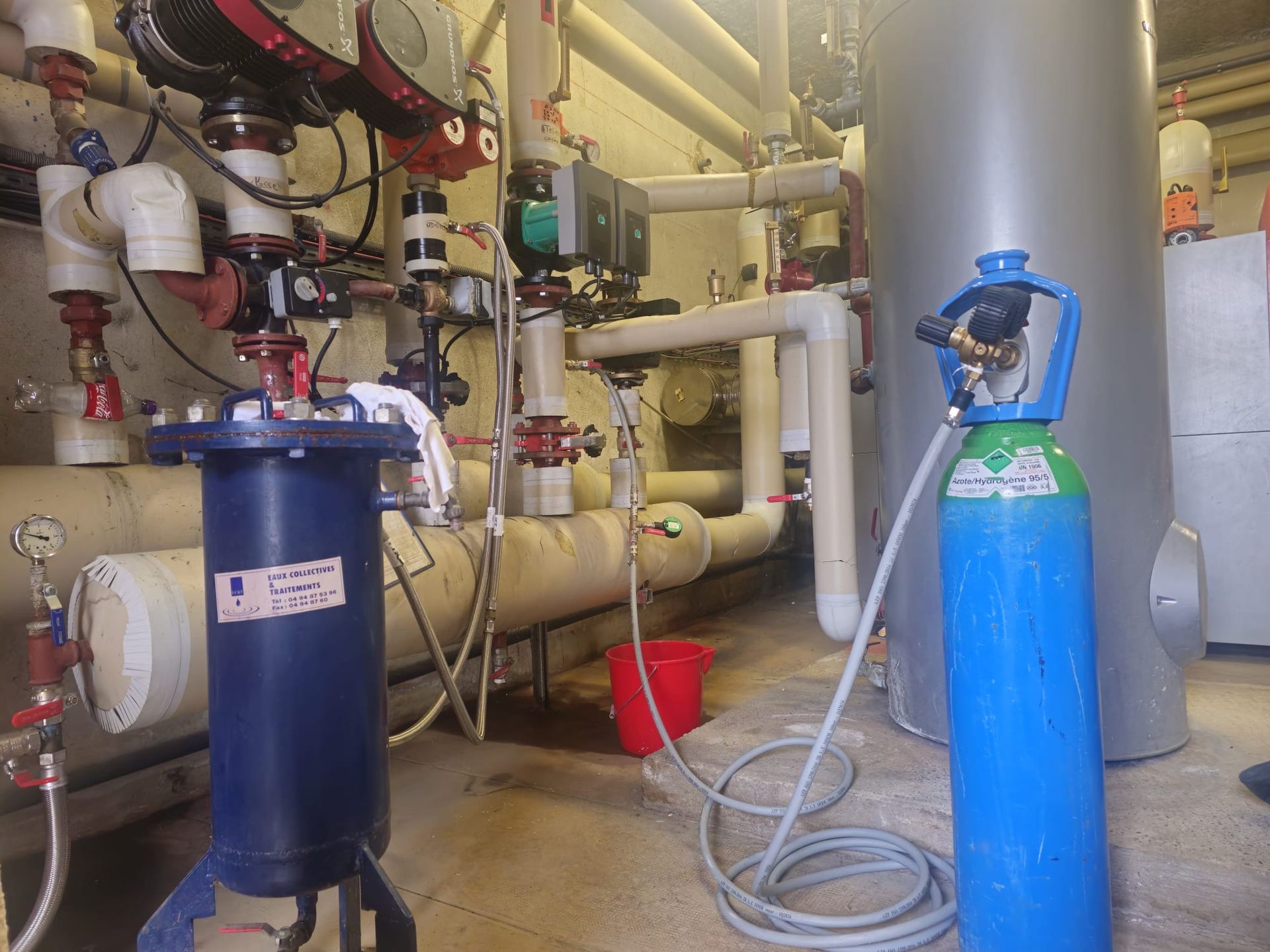 Bouteille de gaz raccordée à des tuyaux dans un local technique où se trouvent d'autres équipements.