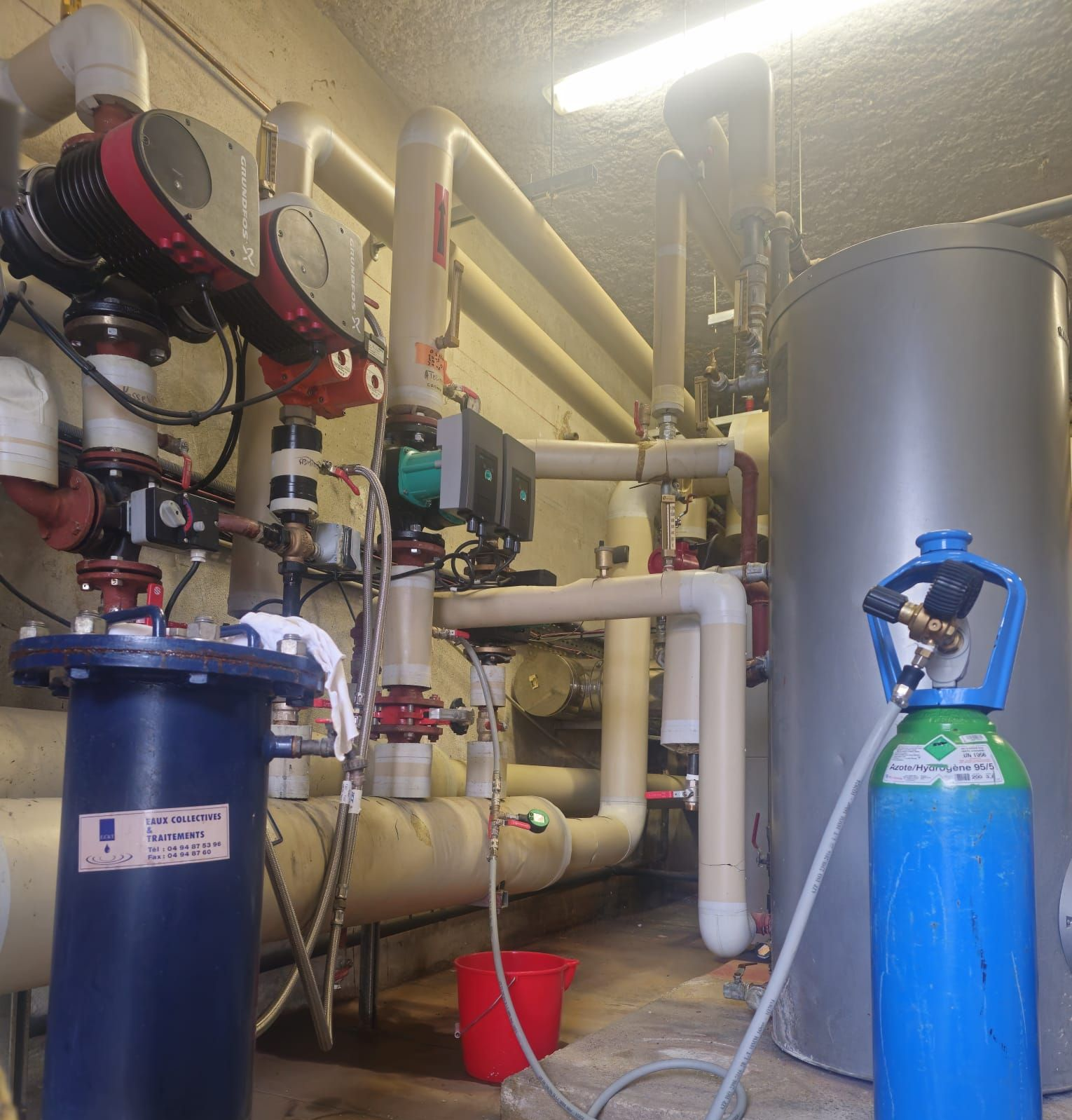 Local industriel avec tuyauterie, vannes, réservoir et équipements. Un seau est posé au sol.