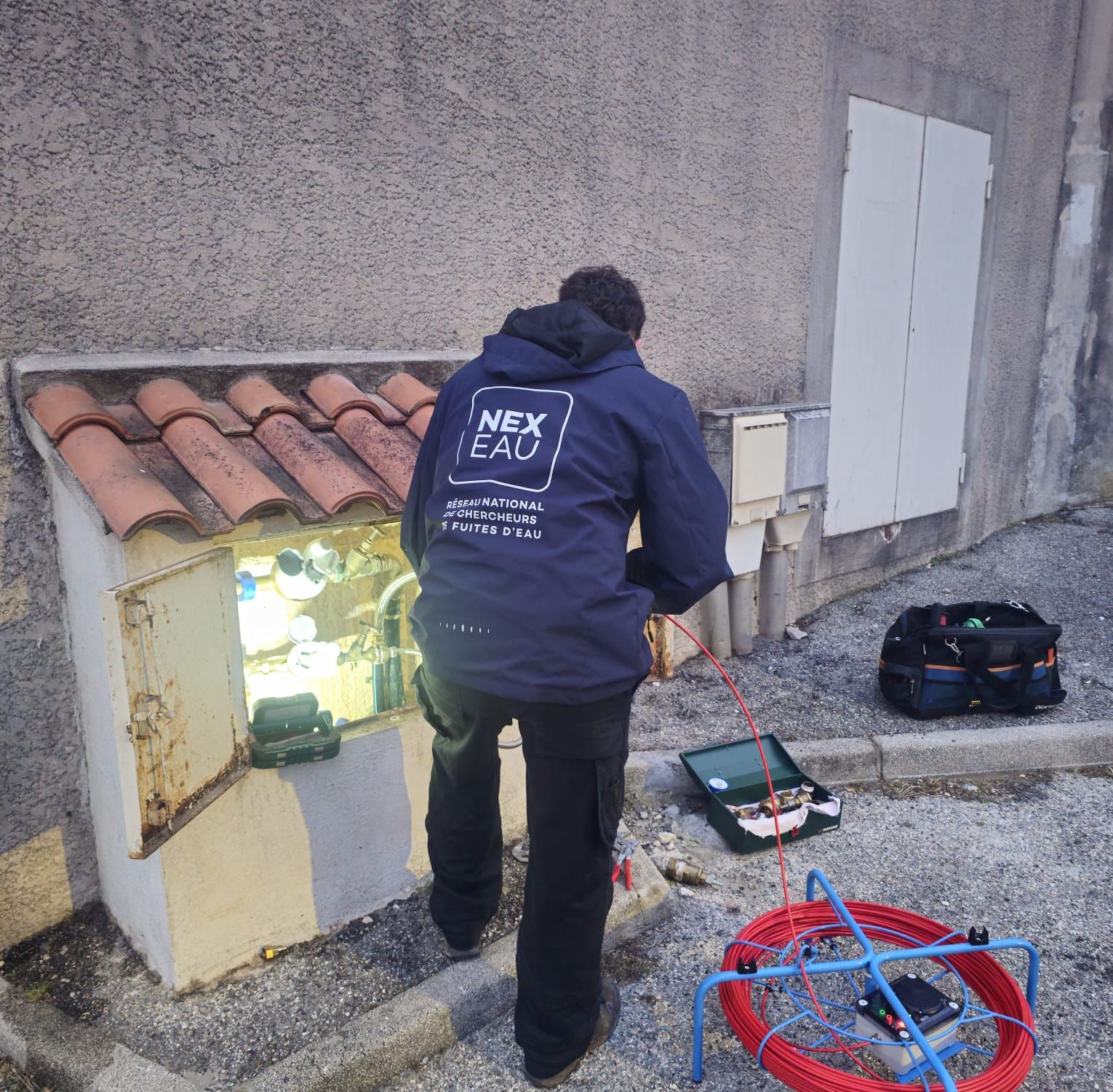 Une personne en veste travaille sur un boîtier de connexion réseau à l'extérieur. Un câble et des outils sont au sol.