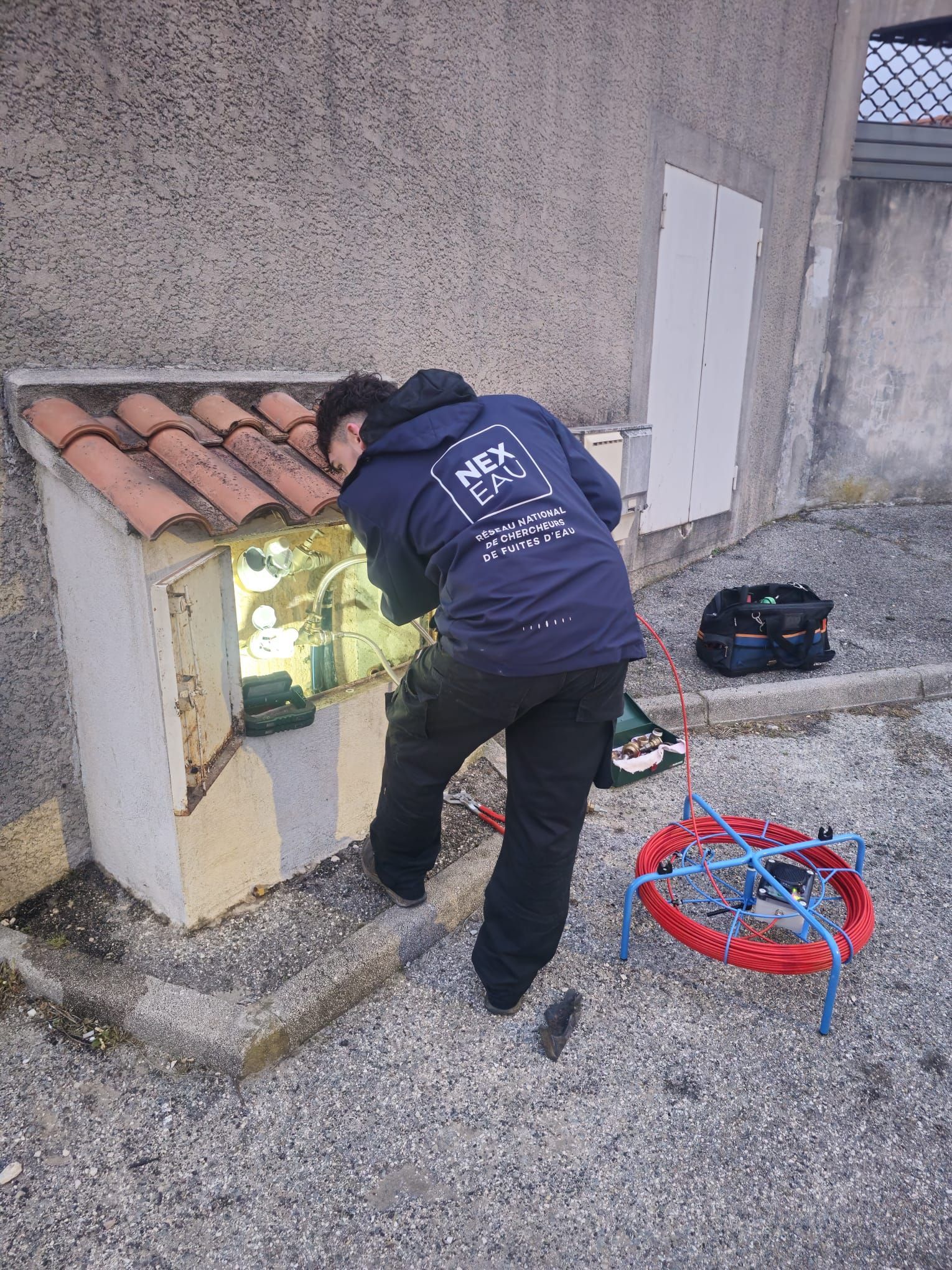 Une personne portant un sweat-shirt à capuche travaille sur le câblage près d'un bâtiment, avec du matériel à proximité.