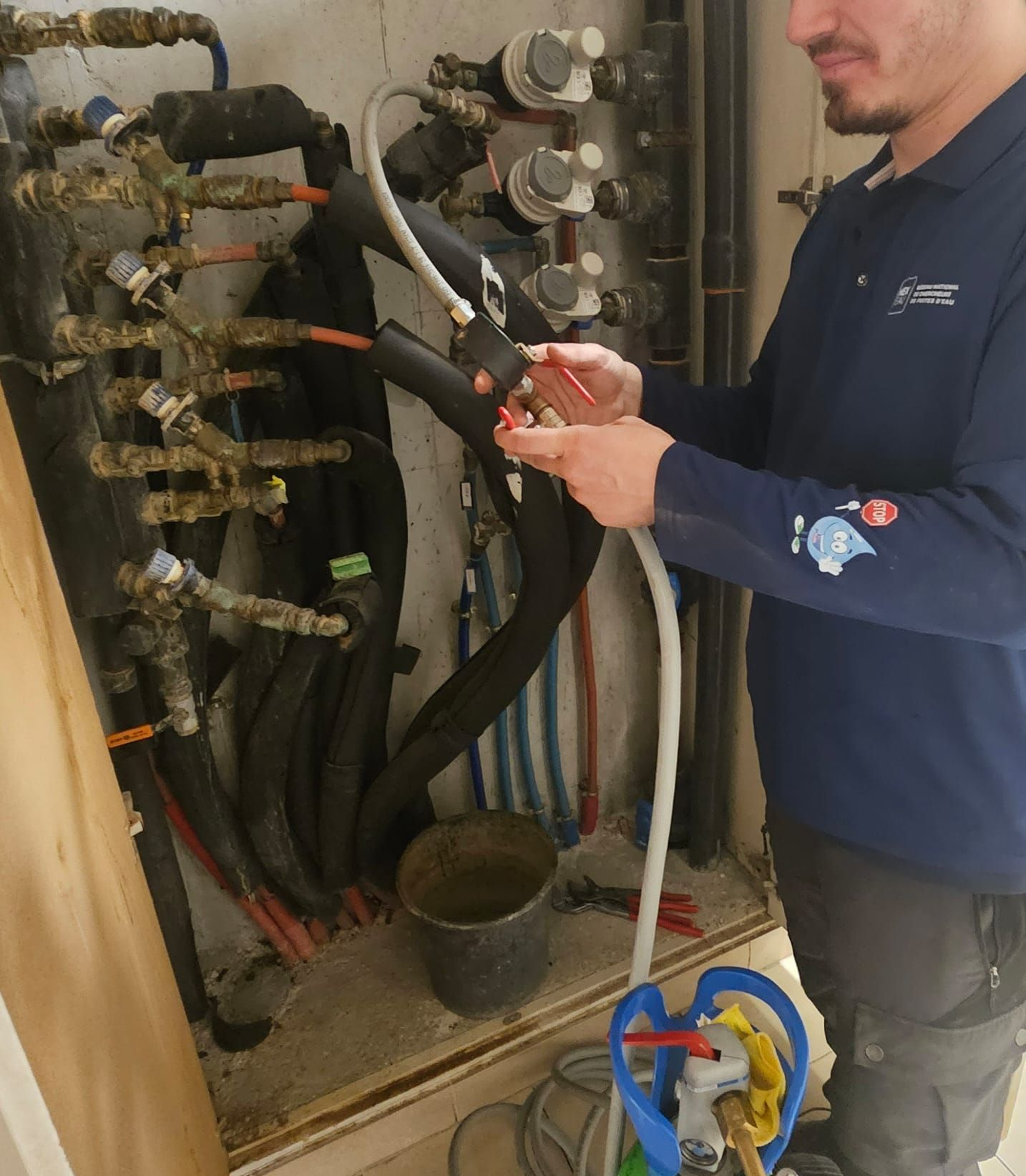 Un plombier travaille sur des tuyaux reliés à un réservoir. Il porte une chemise et un pantalon dans un local technique.