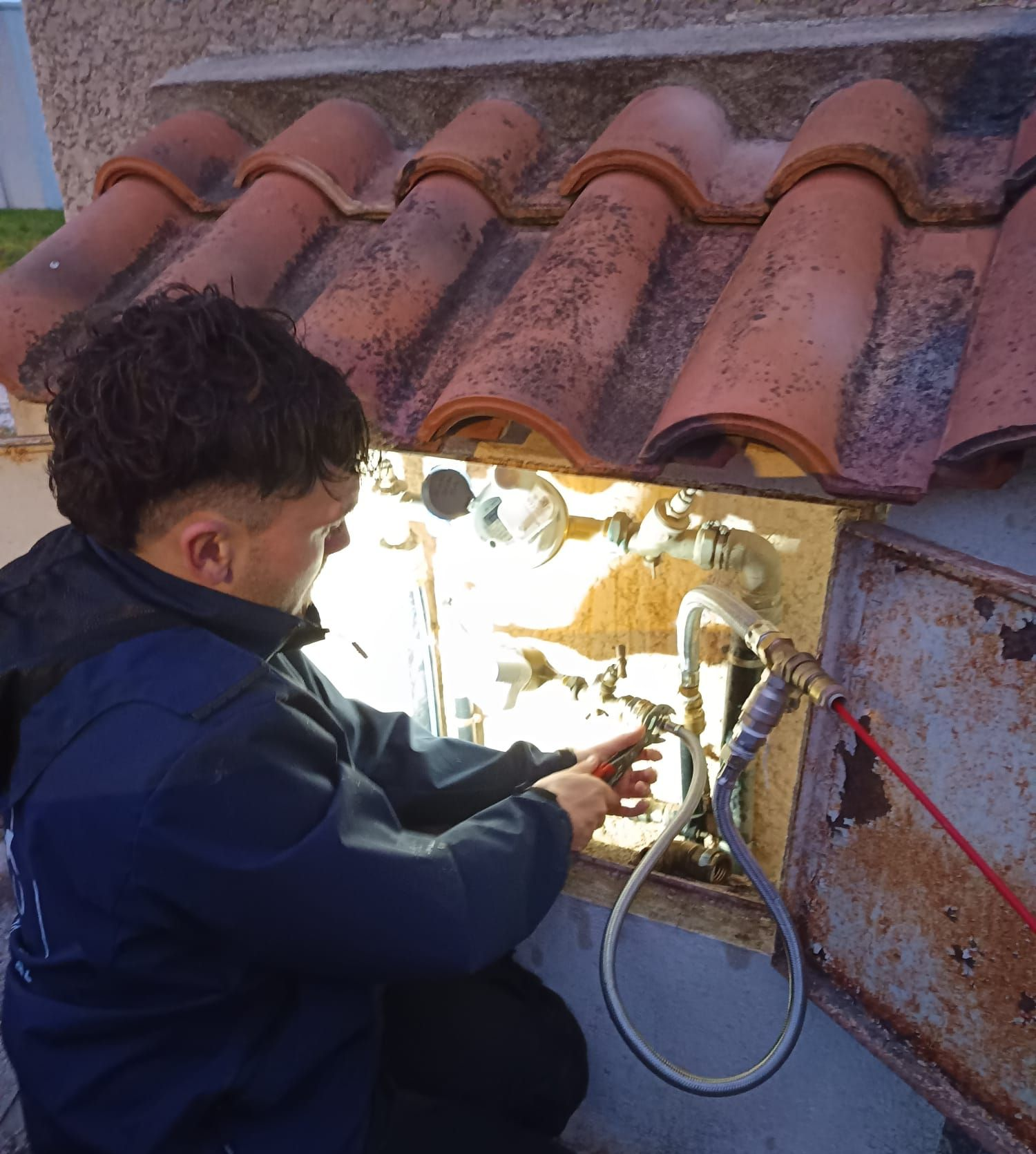 Un plombier travaille sur des canalisations dans une niche sous un toit de tuiles en terre cuite.