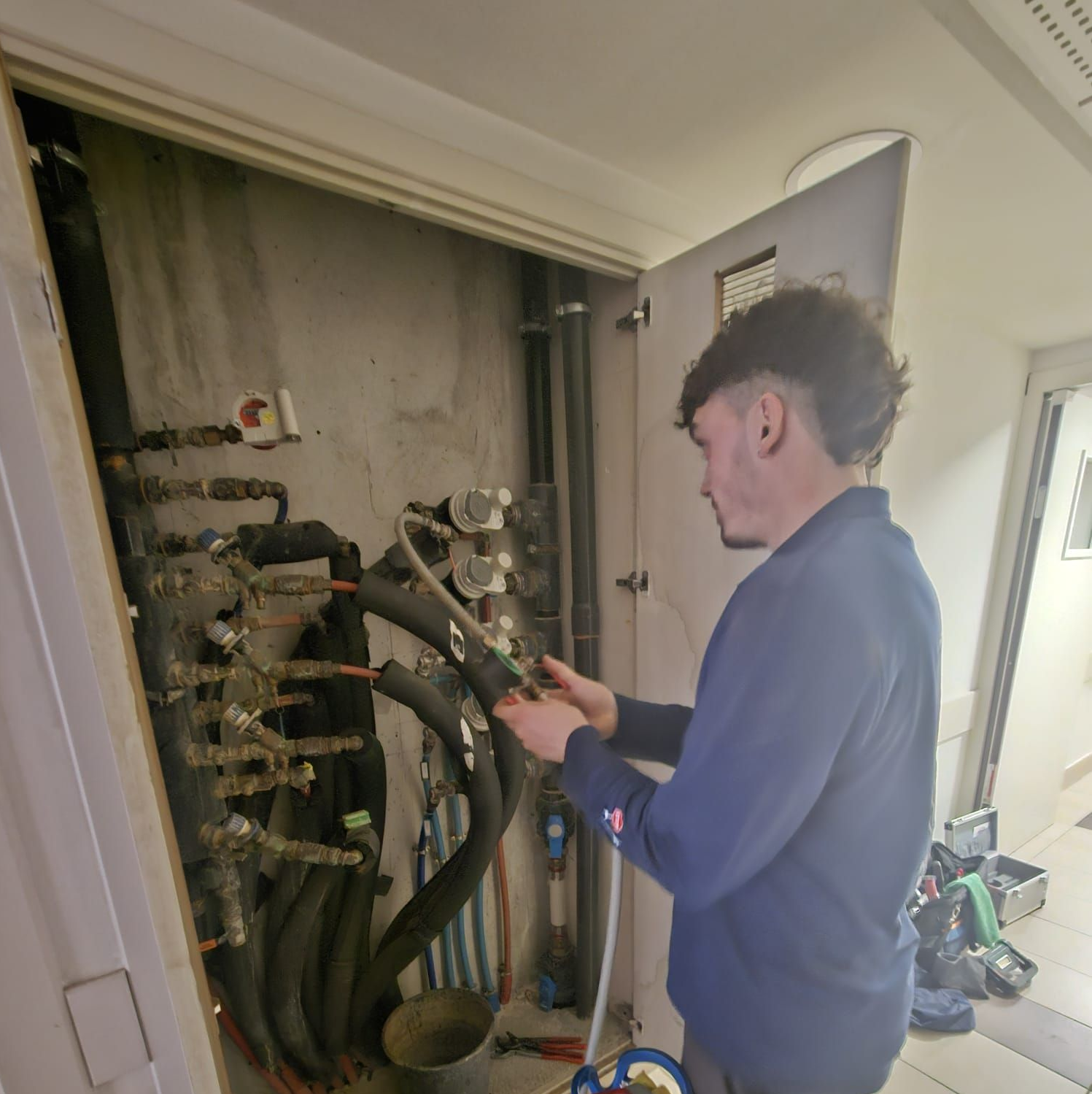 Une personne en chemise travaille sur des tuyaux à l'intérieur d'un local technique, avec un réservoir de gaz à proximité.