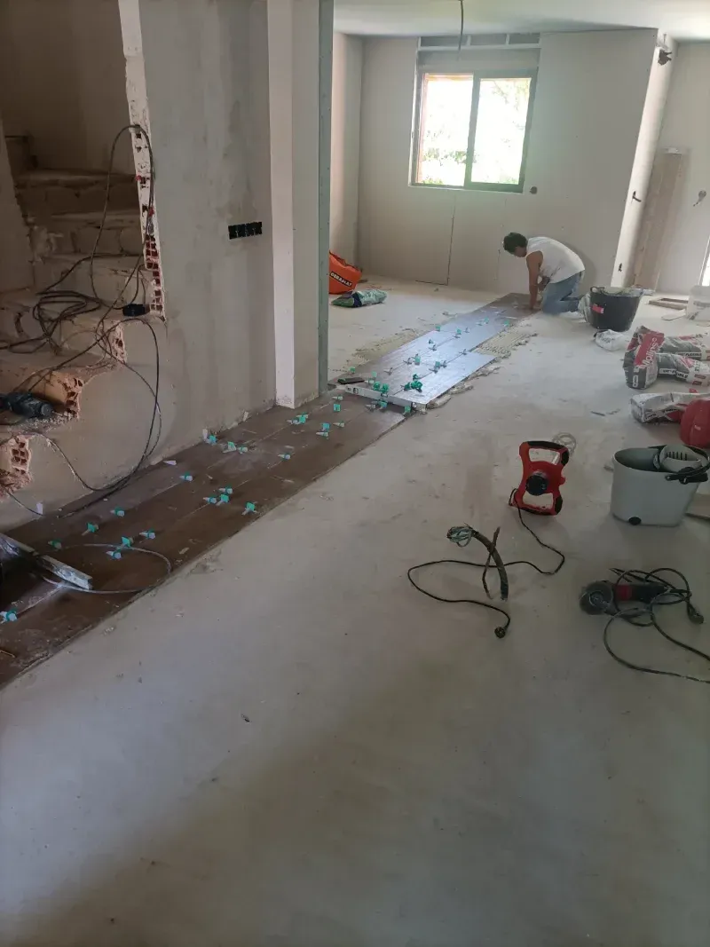 Obra en construcción con una persona colocando suelo; herramientas y materiales esparcidos por el lugar.