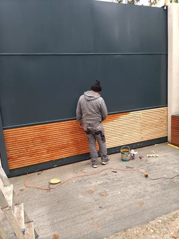Una persona pinta un panel de madera en una gran puerta metálica oscura; hay latas de pintura y herramientas cerca.