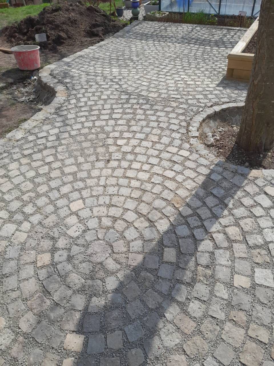 Ein gepflasterter Weg mit geschwungenem Design aus hellbraunen und grauen Steinen führt zu einem Gartenbereich.