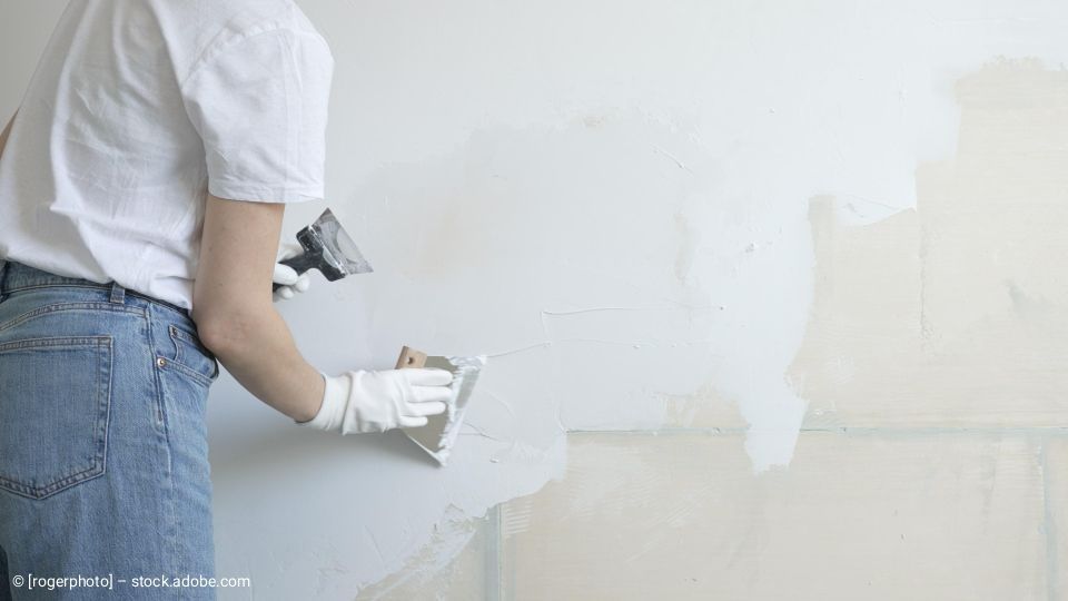 Eine Person, die Handschuhe, Jeans und ein T-Shirt trägt, benutzt eine Kelle, um Gips an eine Wand aufzutragen.