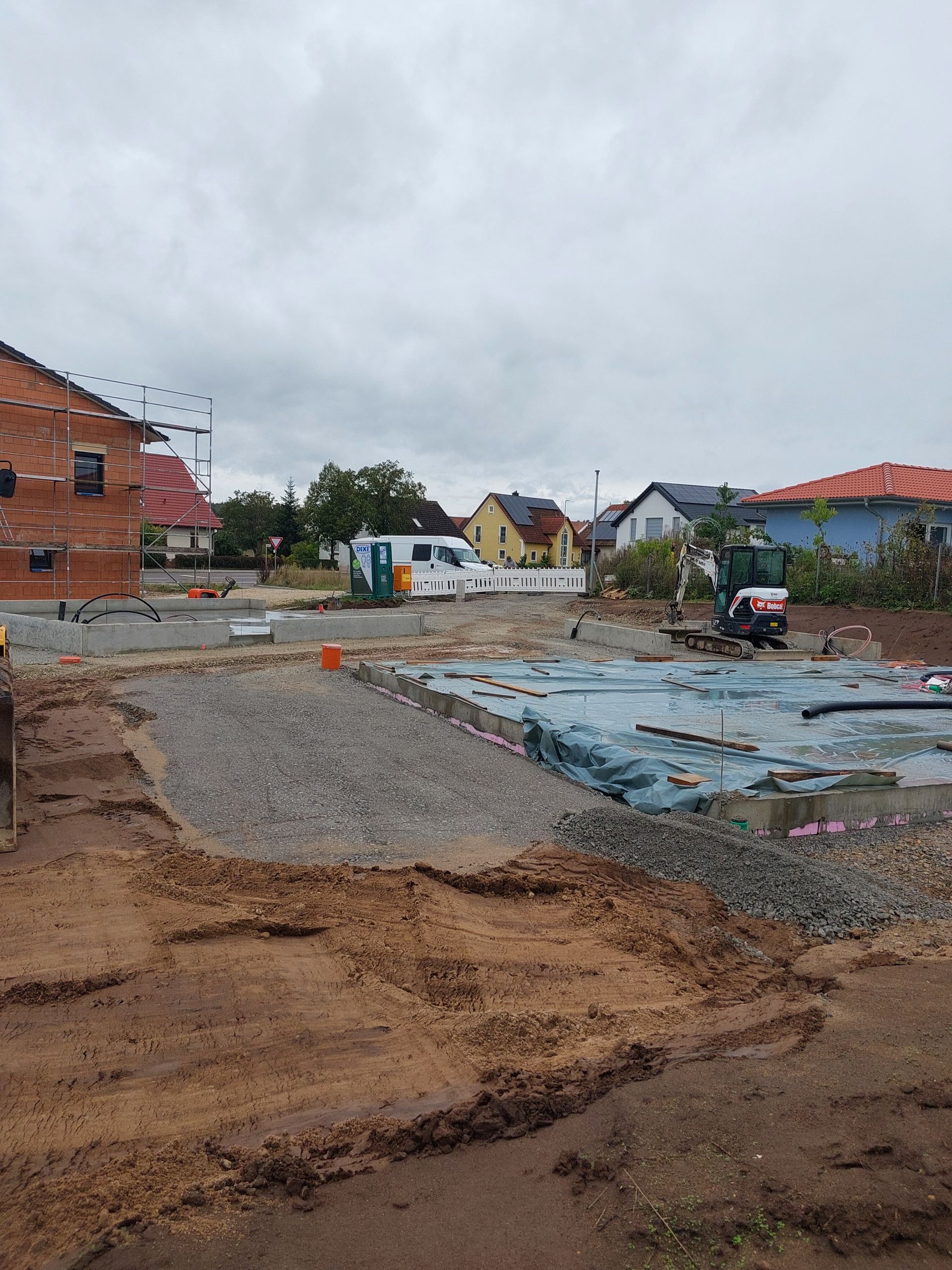 Baustelle mit Betonfundament und Pfeilern, Kies und Kränen im Hintergrund.