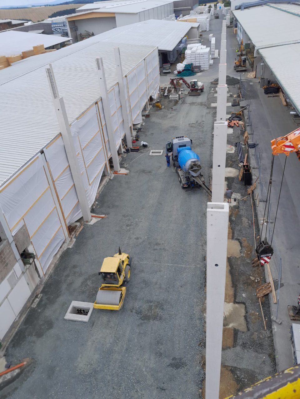 Baustelle mit Betonmischern und einer Walze zur Verdichtung des Kieswegs zwischen zwei langen Gebäuden.