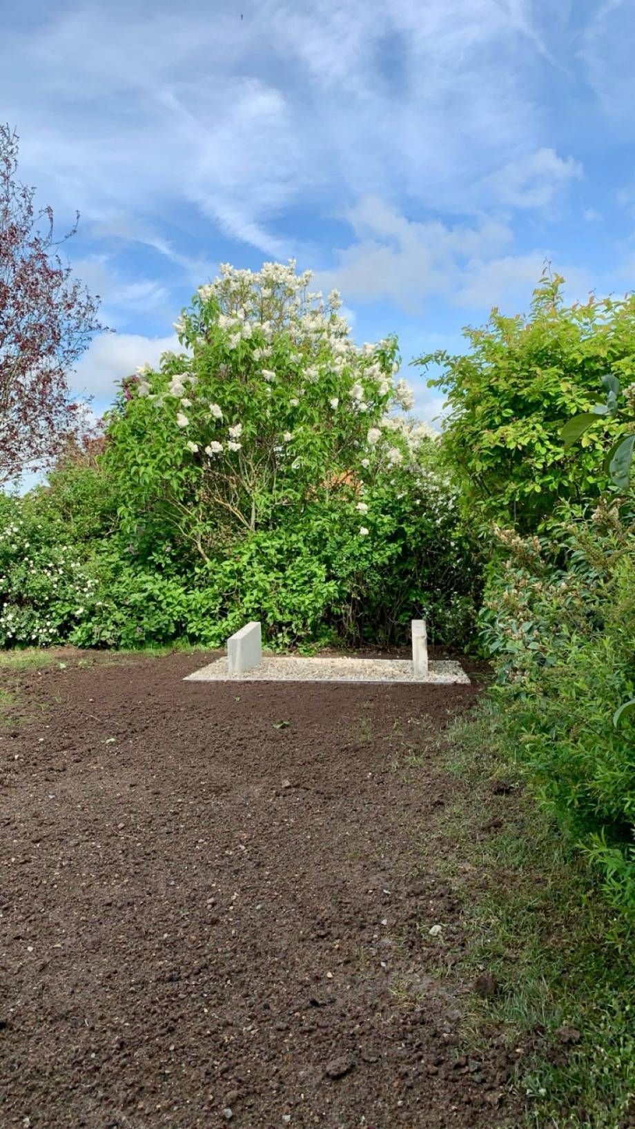 Ein offener Platz mit zwei Steinmarkierungen auf einem Kiesbett, umgeben von grünen Büschen und einem blühenden weißen Baum.