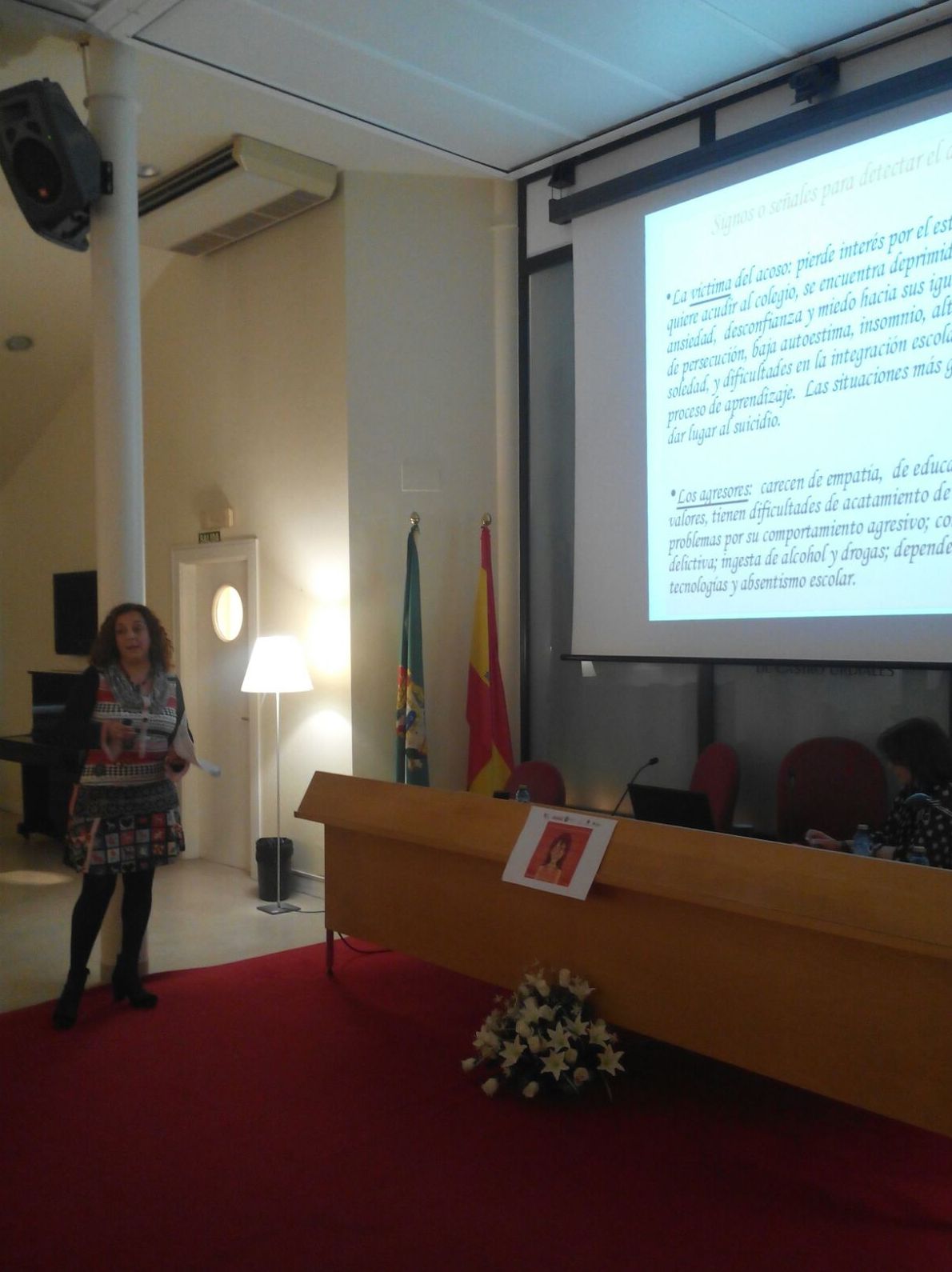 Mujer dando una presentación en un podio con una pantalla que muestra texto en una sala con banderas.