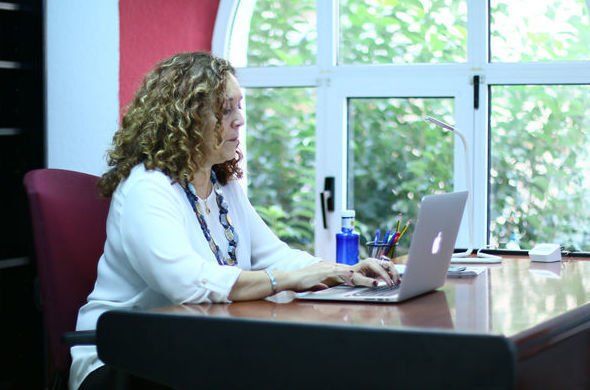 Mujer con cabello rizado escribiendo en una computadora portátil en un escritorio cerca de una ventana, en una oficina.
