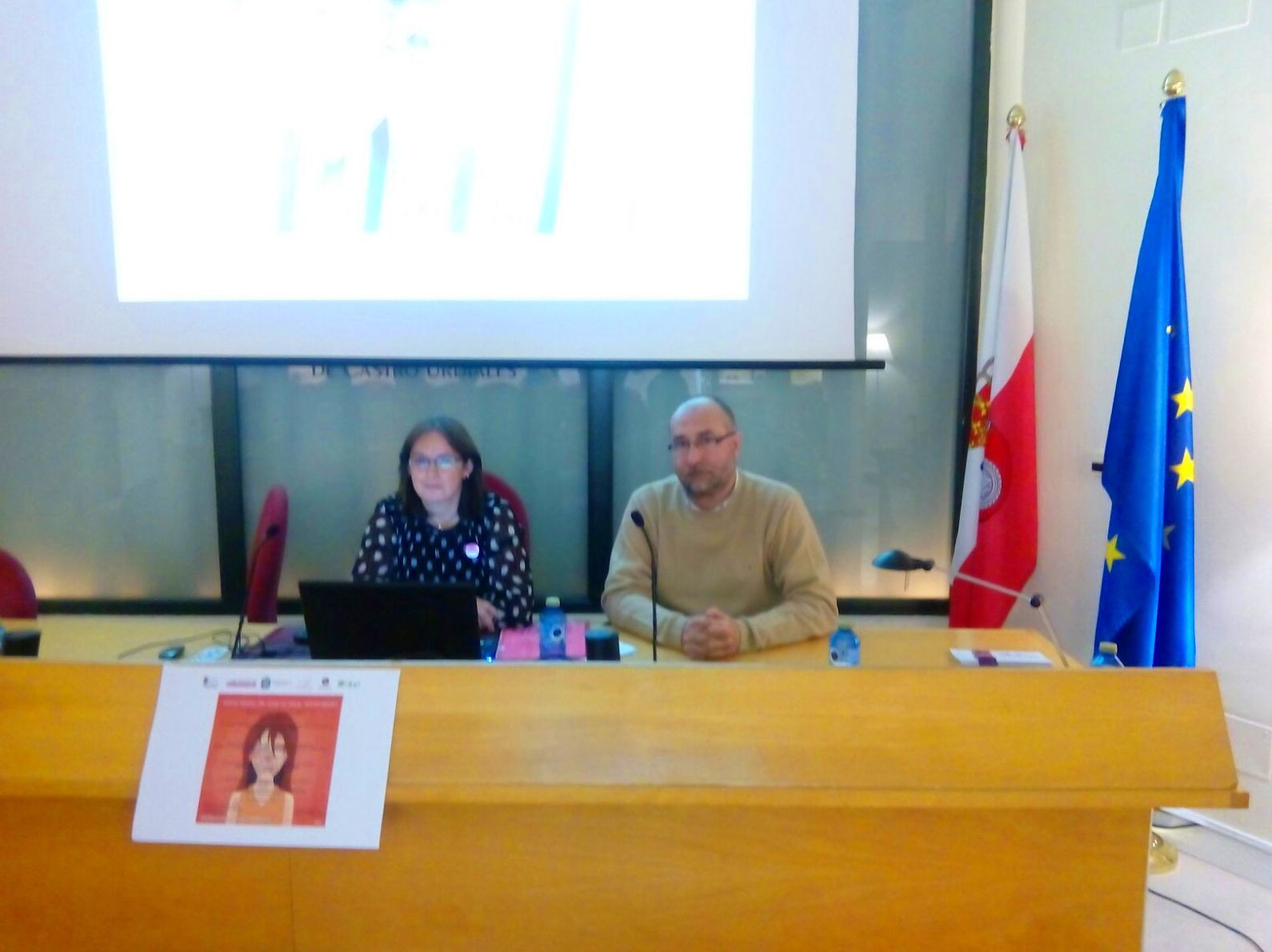Dos personas en una mesa en un entorno de conferencias, una mujer con gafas y un hombre con banderas detrás de ellos.